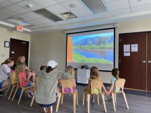 Children in the Coddington Road Community Center's Bear Room are in a classroom looking at a projector screen and are on a virtual train ride.